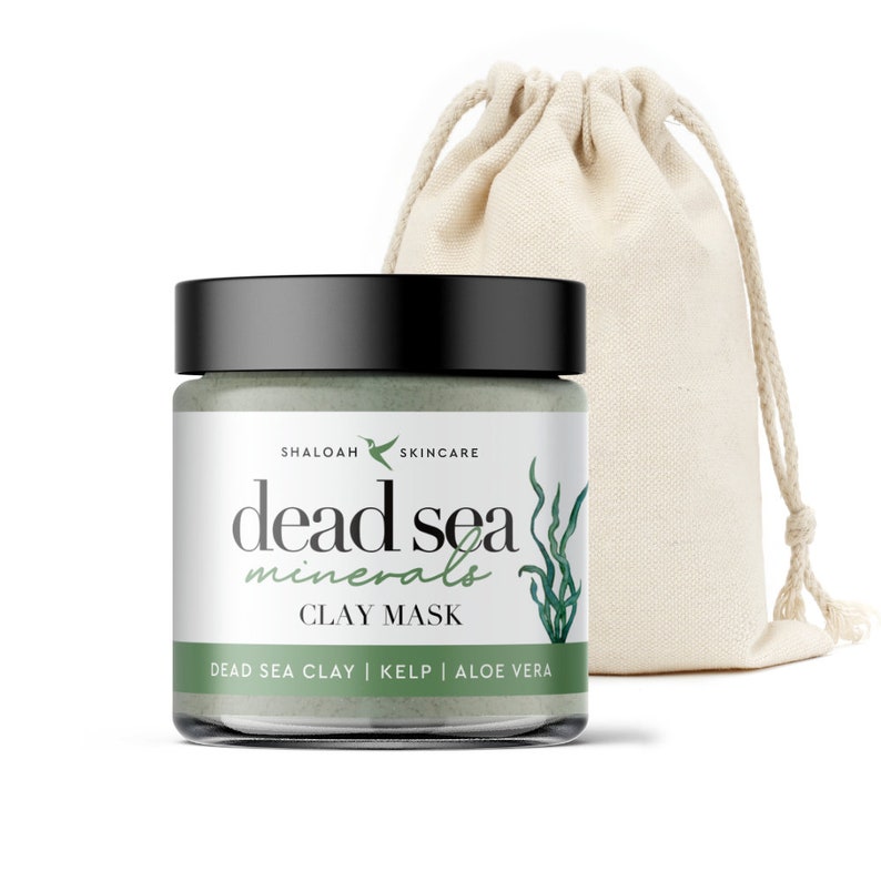 May include: A green glass jar with a black lid containing a Dead Sea Minerals Clay Mask. The jar has a white label with the text "SHALOAH SKINCARE dead sea minerals CLAY MASK DEAD SEA CLAY | KELP | ALOE VERA". The jar is sitting on a white surface next to a white drawstring bag.