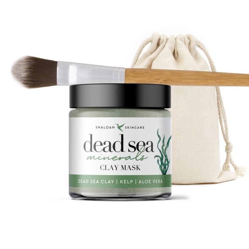 May include: A green glass jar with a black lid containing a Dead Sea Minerals Clay Mask. The jar is labeled with the brand name "Shaloa Skincare" and the ingredients "Dead Sea Clay, Kelp, Aloe Vera". The jar is accompanied by a small white canvas bag and a wooden makeup brush.