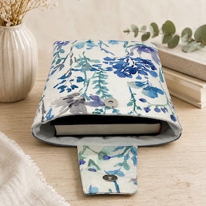 May include: A fabric book sleeve featuring a watercolour floral pattern in shades of blue, teal, and grey on a white background. The sleeve has a fold-over flap with a silver-toned magnetic snap closure and contains a black-bound book inside.