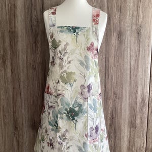 May include: A white apron with a floral print in shades of green, purple, and pink. The apron has two pockets and adjustable straps.