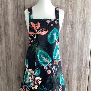 May include: Black apron with a floral print featuring pink, green and orange leaves and flowers. The apron has two pockets and adjustable straps.