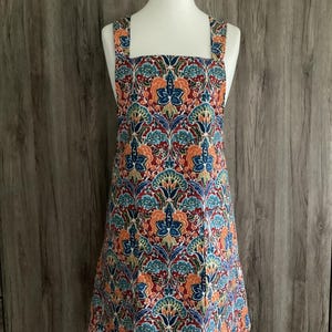 Puede incluir: Delantal colorido con un estampado floral en tonos rojos, azules y naranjas. El delantal tiene un escote cuadrado y tirantes anchos. La tela está adornada con flores y hojas estilizadas, creando una estética vintage.