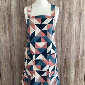 May include: An apron with a geometric pattern of triangles in shades of blue, pink, and white. The apron has two front pockets and a cross-back design. The apron is displayed on a mannequin.