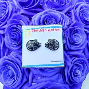 May include: A pair of hypoallergenic earrings featuring a black and white image of the Millennium Falcon from Star Wars. The earrings are displayed on a white card with the text "Childish Antics" and "Hypoallergenic".