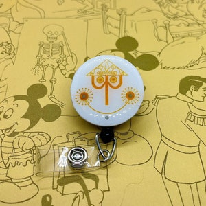 May include: A white and orange round badge holder with a stylized tree design and three yellow flowers. The badge holder is attached to a clear plastic retractable badge reel with a black clip.