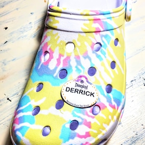 May include: A tie-dye Croc shoe with a white interior and a Disneyland name tag that reads "Derrick" and "Bringing Back The Magic". The shoe features a pattern of yellow, pink, blue, and white swirls.