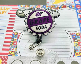 May include: A purple and white grape soda bottle cap badge reel with a safety pin and a retractable badge holder. The badge reel features the text "GRAPE SODA" and a bunch of grapes.
