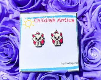 May include: A pair of hypoallergenic stud earrings featuring a cartoon image of a white cat with pink accents. The earrings are displayed on a white card with the text "Childish Antics" and "Hypoallergenic".