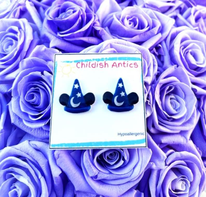 Op de afbeelding: Een paar blauwe en zilveren Mickey Mouse-oren met een ster- en maanontwerp. De oorbellen zijn hypoallergeen en worden gepresenteerd op een witte kaart met de tekst "Childish Antics".