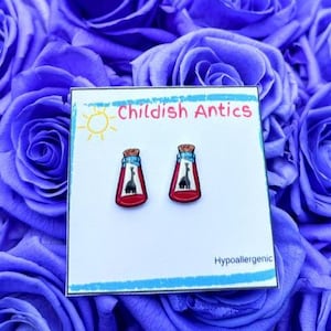 May include: A pair of red and clear bottle-shaped earrings with a giraffe silhouette inside, displayed on a white card with the text "Childish Antics" and "Hypoallergenic". The earrings are set against a backdrop of blue roses.