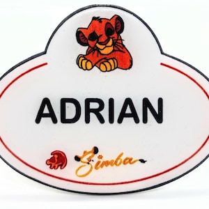 May include: A white name tag with a red border and the name "ADRIAN" in bold black letters. A cartoon lion cub is at the top, and the name "Simba" is written in cursive below.