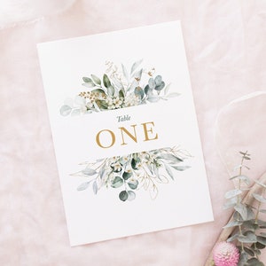 May include: White card with a gold floral design and the text "Table ONE" in gold lettering.