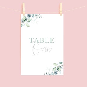 May include: A white table number card with the text "TABLE One" in a soft, elegant font. The card is adorned with watercolor eucalyptus leaves in shades of green and blue. The card is clipped to a string with wooden clothespins against a pink background.