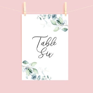May include: White card with green eucalyptus leaves printed on the top and bottom. The card says "Table Six" in black cursive font.