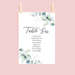 May include: A white card with the text "Table Six" in elegant script, listing names below. The card is decorated with watercolor eucalyptus leaves and is suspended by a wooden clothespin against a pink background.