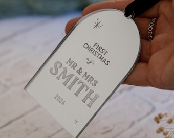 May include: A silver-colored Christmas ornament with rounded top edges and a black ribbon. The ornament is engraved with "FIRST CHRISTMAS", "MR & MRS SMITH", and "2024". A star and heart are also engraved on the ornament.