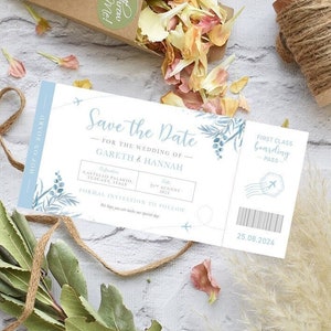 May include: A blue and white save the date card with a plane ticket design. The card says "Save the Date" for the wedding of Gareth & Hannah on 25th August 2024. The card is surrounded by dried flowers and greenery.