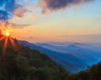 Puede incluir: Una vista panorámica de una cordillera al amanecer con el sol brillando a través de las nubes e iluminando las laderas boscosas.