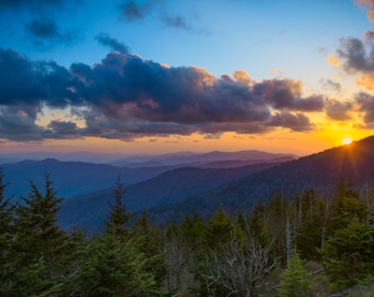 May include: A scenic view of a mountain range at sunset. The sun is setting behind the mountains, casting a warm glow on the landscape. The sky is a mix of blue, orange, and purple, and there are clouds in the sky. The mountains are covered in trees, and there is a forest in the foreground.