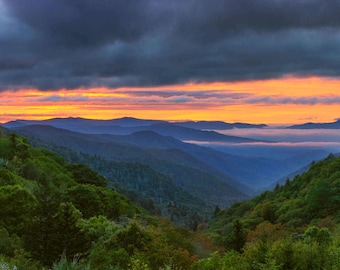 May include: A panoramic view of a mountain range at sunrise. The sky is a vibrant orange and pink, with dark clouds in the distance. The mountains are covered in lush green trees.