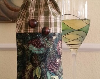 May include: A fabric wine bottle bag with a green plaid top and a forest-themed bottom. The bag is tied with a twine and has two wooden beads. A wine glass with a green and black design is next to the bag.