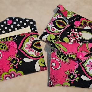 May include: Three pink, green, and black fabric pouches with pink buttons. The pouches are made of a patterned fabric with a paisley design. The pouches are folded in half and have a flap that closes with a button.