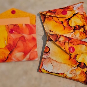 May include: Three fabric envelopes with a yellow and orange marbled pattern and pink buttons. The envelopes are folded and have a flap on the top.