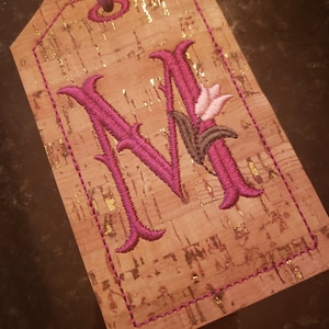 May include: A cork luggage tag with a pink and purple embroidered letter "M" with a pink flower and green leaves.