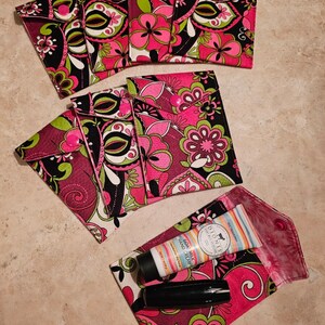 May include: A set of eight pink, green, and black fabric pouches with a button closure. The pouches are decorated with a paisley pattern. A small tube of hand cream and a black lipstick are inside one of the pouches.