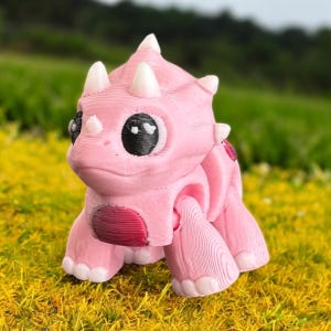 Puede incluir: Figura de Triceratops rosa impresa en 3D con cuernos blancos y grandes ojos negros. El dinosaurio tiene una expresión amigable y está posado sobre hierba amarilla. La figura tiene una superficie detallada y texturizada.