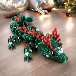 May include: A green and red decorative dragon figurine with a velvet texture. The dragon has red accents on its back and head, with white pearl-like embellishments. The dragon is lying on a wooden surface, with Christmas decorations in the background.