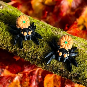 Puede incluir: Dos figuras de arañas negras con detalles naranjas y pequeñas calabazas encima. Las arañas están posadas en una rama cubierta de musgo, con hojas de otoño de fondo. Una decoración festiva de Halloween.