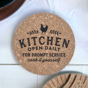 Puede incluir: Posavasos de cocina redondo de corcho con una ilustración de gallo negro y las palabras "KITCHEN OPEN DAILY FOR PROMPT SERVICE cook it yourself". El posavasos está hecho de corcho natural y protege las superficies del calor.