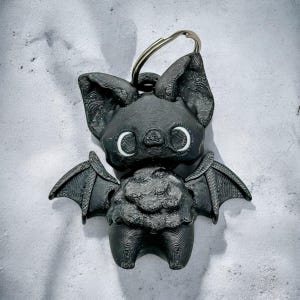 May include: A black bat-shaped keychain with a silver keyring. The bat has large ears, wings, and white eyes. The bat's body has a textured, layered appearance, and the background is a light gray.