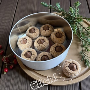 May include: A white tin filled with peanut butter cookies with a chocolate center. The cookies are on a wooden tray with a sprig of greenery. The word "Click!" is written in white on the bottom right corner.