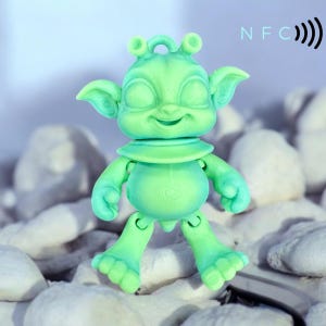 May include: A light green alien figurine with large ears, a smiling face, and a collar. The figurine has articulated arms and legs. The letters "NFC" and a radio wave symbol are visible in the background. The figurine is standing on white stones.