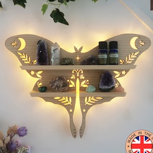 May include: Illuminated wooden wall shelf shaped like a moth with two shelves.  The shelf features cut-out floral designs and moons. It displays crystals, essential oil bottles, and other small objects.  The shelf is light brown and has a warm, ambient light.  A label reads 'MADE IN THE UK'.