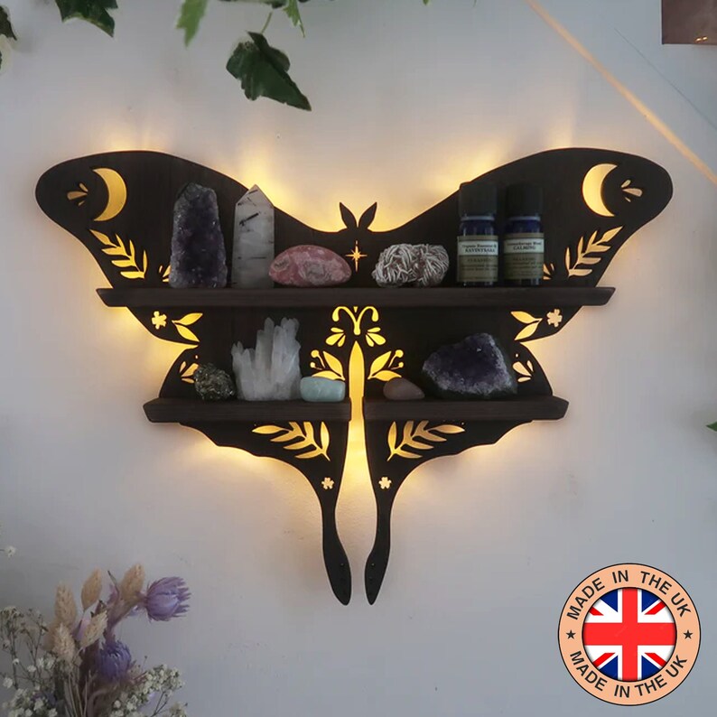 May include: Illuminated dark brown wooden butterfly-shaped crystal and essential oil shelf; two tiers with cut-out floral details and crescent moons; holds various crystals, including amethyst, clear quartz, and selenite; two small essential oil bottles are on the top shelf;  'MADE IN THE UK' label is visible.