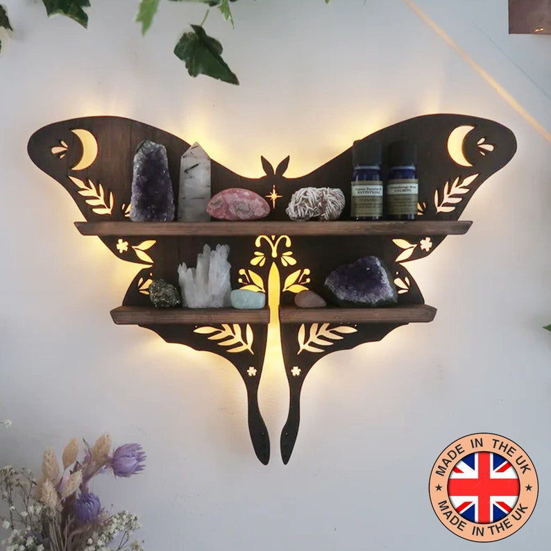 May include: Dark brown wooden butterfly-shaped shelf with two tiers, illuminated from behind.  Intricate cut-out designs feature leaves, flowers, and moons. The shelves display various crystals, tumbled stones, and small bottles, suggesting a crystal or essential oil organizer.  A label reads 'MADE IN THE UK' with a UK flag graphic.