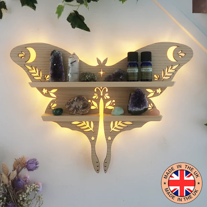 May include: Illuminated wooden wall shelf shaped like a moth with two shelves.  The shelf features cut-out floral designs and moons. It displays crystals, essential oil bottles, and other small objects.  The shelf is light brown and has a warm, ambient light.  A label reads 'MADE IN THE UK'.