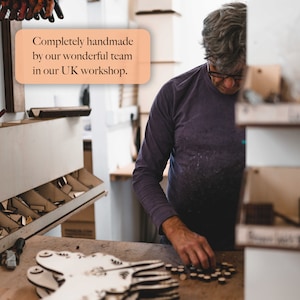 May include: Wooden crafts are being assembled in a UK workshop.  A craftsman carefully places small wooden pieces onto a larger unfinished wooden cutout. The scene is warm and inviting, showcasing the handmade nature of the products. Text overlay reads: 'Completely handmade by our wonderful team in our UK workshop.'