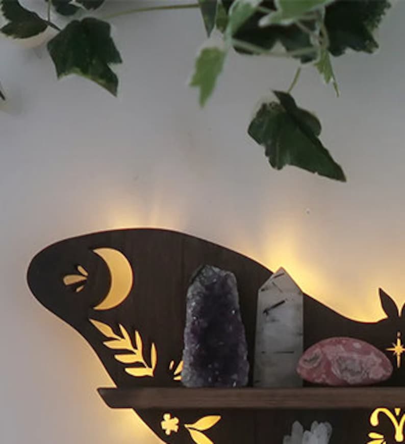 May include: Dark brown wooden crystal shelf with LED lights, featuring cut-out moon, stars, and floral designs.  Amethyst, clear quartz, and rose quartz crystals are displayed on the shelf. The shelf has a whimsical, celestial theme, perfect for showcasing crystals and minerals.  It's a wall-mounted shelf with a unique shape and intricate detailing.