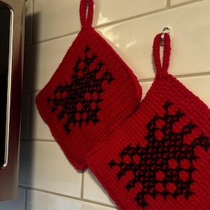 Könnte beinhalten: Zwei rote gehäkelte Topflappen mit schwarzen geometrischen Mustern. Die Topflappen haben Schlaufen zum Aufhängen.