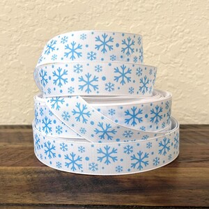 May include: A spool of white grosgrain ribbon featuring a repeating pattern of light blue snowflakes and small dots. The ribbon is wound into a cylindrical shape, displaying the textured fabric surface and the consistent winter-themed print.