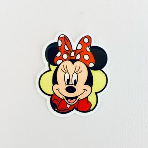May include: A red and white polka dot bow sits on top of Minnie Mouse's head. She is wearing a red dress and has a yellow background.