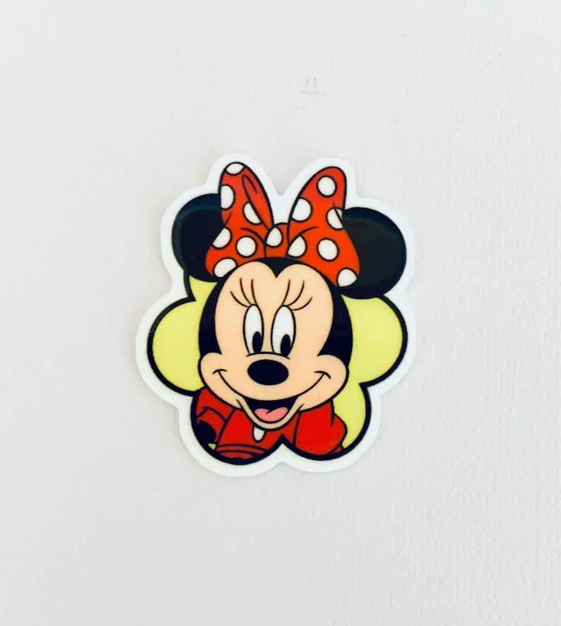 May include: A red and white polka dot bow sits on top of Minnie Mouse's head. She is wearing a red dress and has a yellow background.