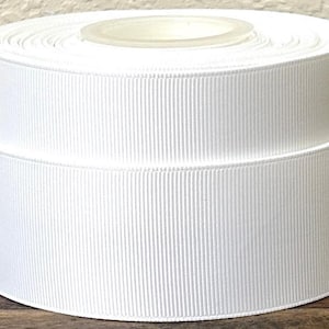 May include: A roll of white grosgrain ribbon. The ribbon is tightly wound and has a ribbed texture. The spool is made of a clear plastic material. The ribbon is sitting on a wooden surface.