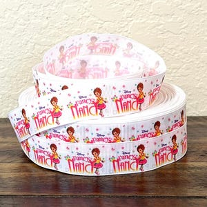May include: White ribbon featuring the Disney character Fancy Nancy. The ribbon has a repeating pattern of Nancy in a yellow top and pink skirt, with the words "Fancy Nancy" and small floral accents. The ribbon is rolled up on a wooden surface.
