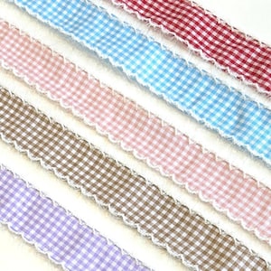 May include: Five strips of fabric ribbon arranged diagonally, each featuring a small gingham check pattern in red, light blue, pink, brown, and purple. Each ribbon has a frayed, raw white edge along both sides of the length of the strip.