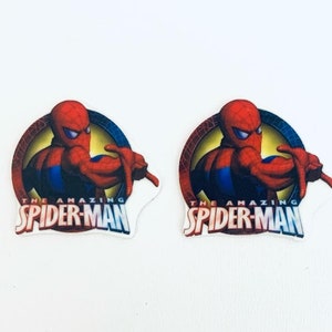 以下が含まれることがあります： アメージング・スパイダーマンのロゴが描かれた、赤、青、黄色の丸いステッカー2枚。ロゴには、赤いスーツと青いスーツを着たスパイダーマンの漫画が描かれており、ウェブデザインが施されています。テキスト「The Amazing Spider-Man」は白い文字で書かれています。